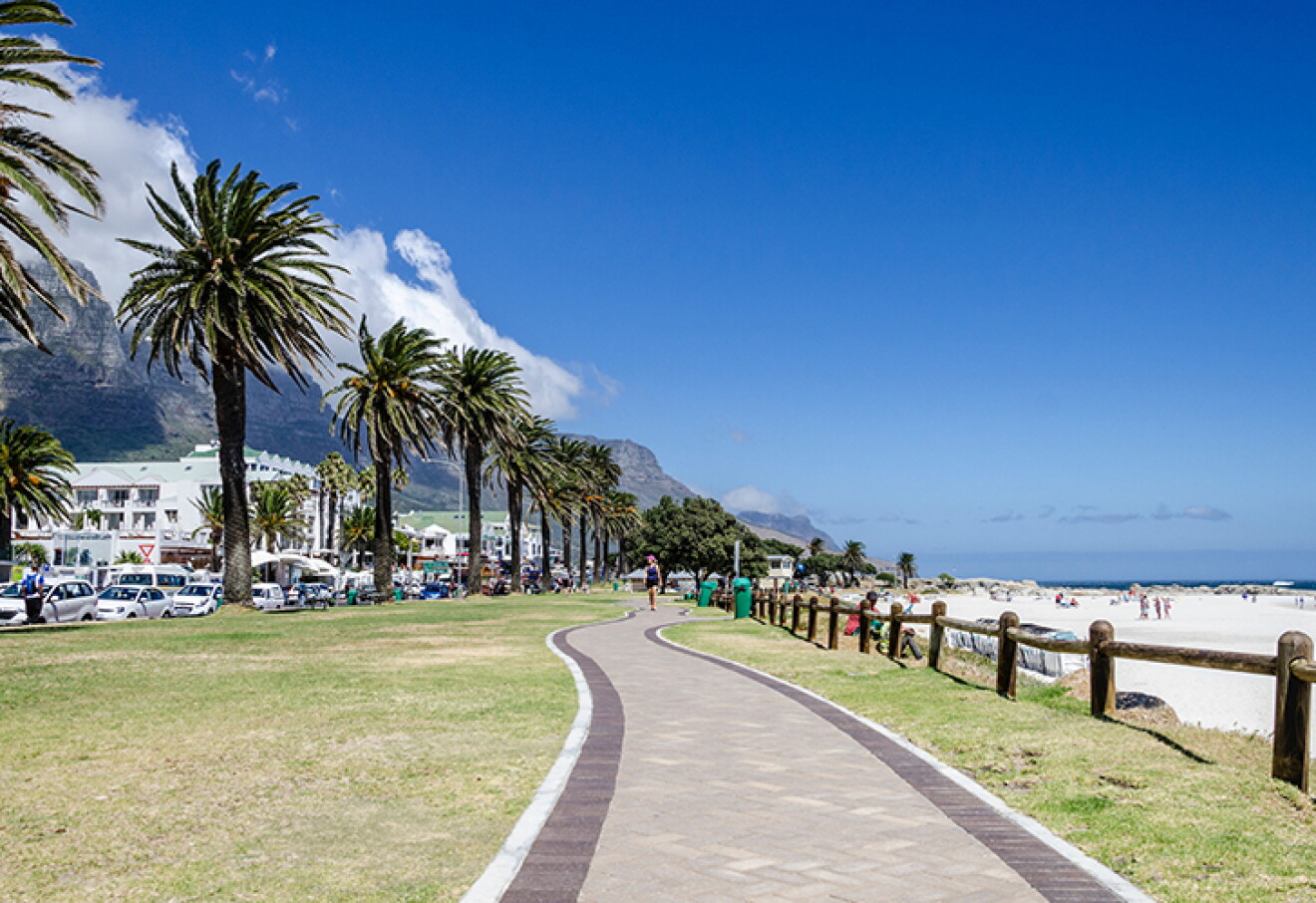 A paved promenade lined with tall palm trees runs alongside a crowded sandy beach with people enjoying the sun, while a mountain range and several buildings are visible in the background under a bright blue sky.