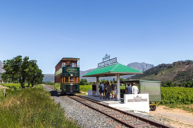 A group of people wait at a small station with a green-roofed shelter beside a vintage double-decker tram labeled "Franschhoek Wine Tram," set against a backdrop of lush vineyards and distant mountains under a clear blue sky.