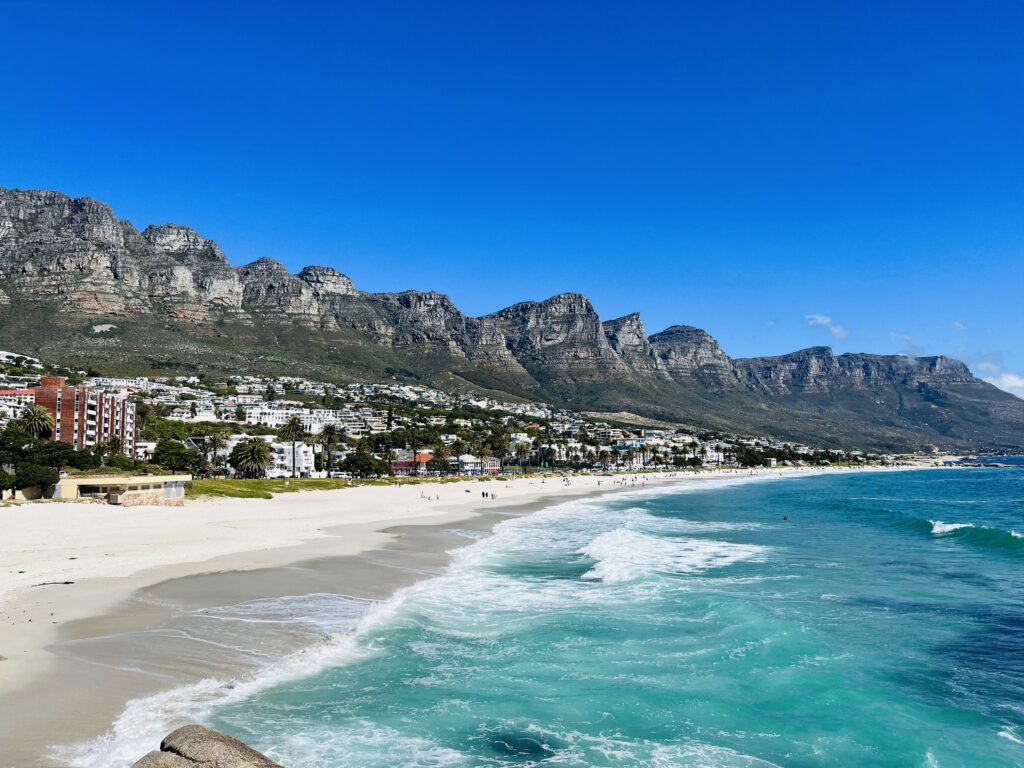 A scenic view of a pristine beach with white sand, turquoise waves, lush green palm trees, and a line of buildings at the foot of rugged mountains under a clear blue sky.