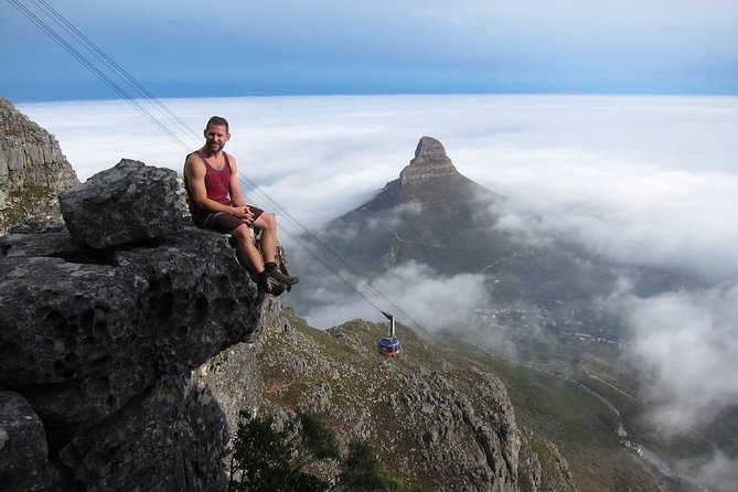 A person sits on a large rock near the edge of a mountain with a view of Table Mountain and its cable car, surrounded by clouds and mist over the landscape.
