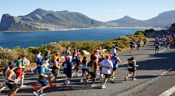 A large group of runners participating in a marathon along a coastal road with mountains and a body of water in the background, under a clear blue sky.