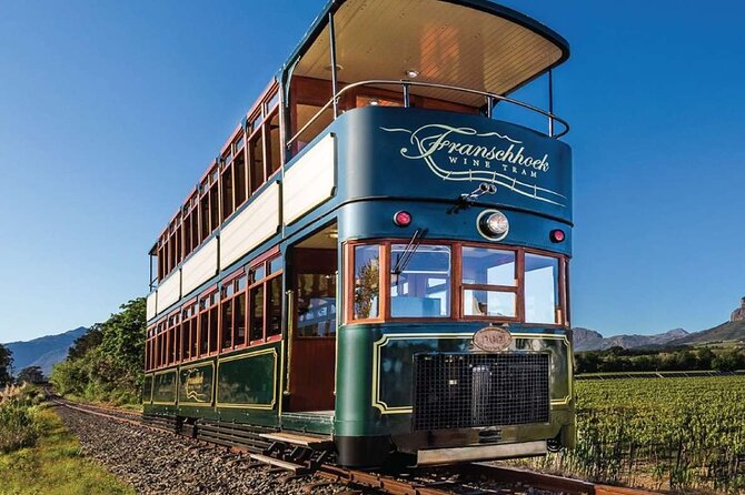 A vintage double-decker tram painted in green and blue with "Franschhoek Wine Tram" branding on the front, traveling along a railway track through lush vineyards against a backdrop of mountains under a clear blue sky.