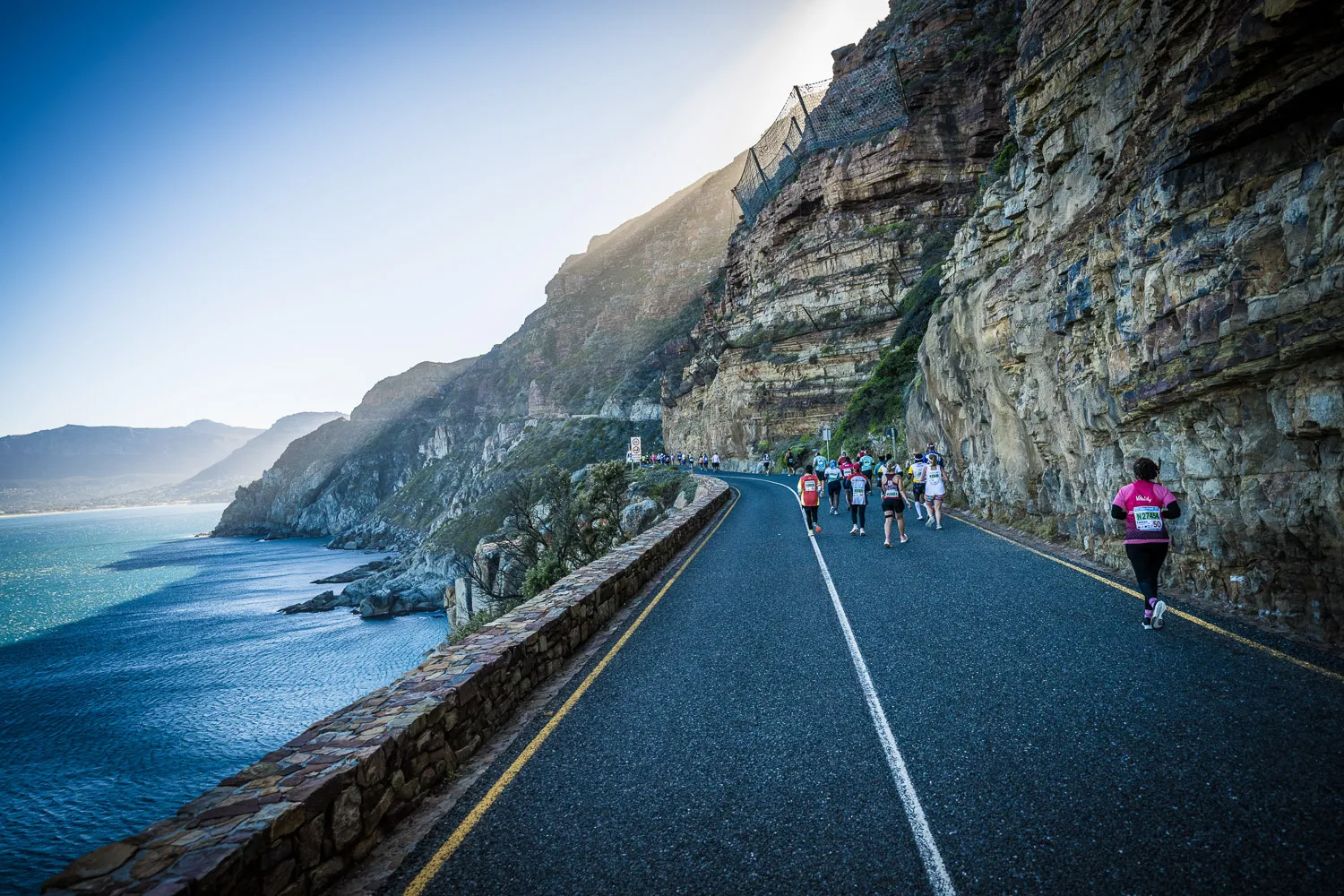 A group of marathon runners participate in the Two Oceans Marathon along a scenic coastal road with steep cliffs and the ocean on one side, under clear skies in Cape Town, South Africa.